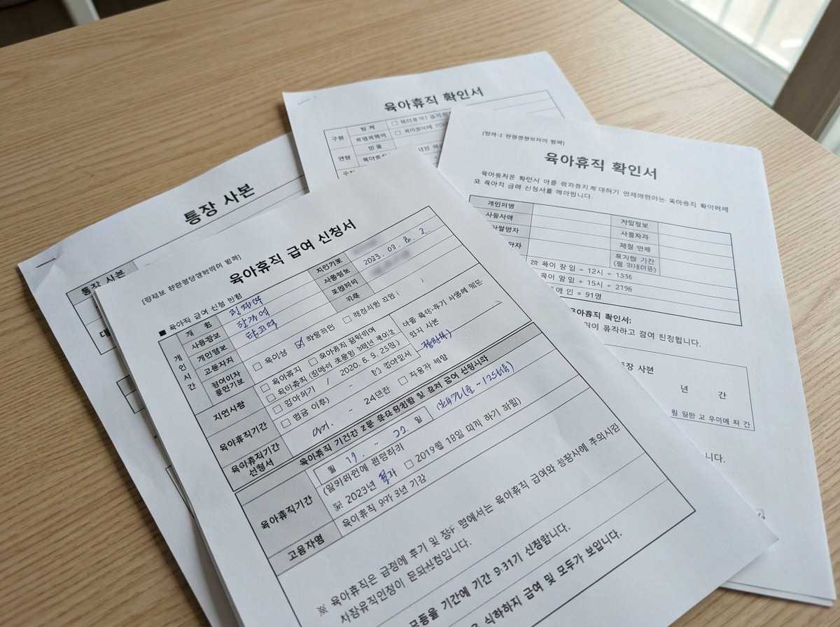 테이블 위에 놓인 육아휴직 급여 신청서, 육아휴직 확인서, 통장 사본 등 서류 더미